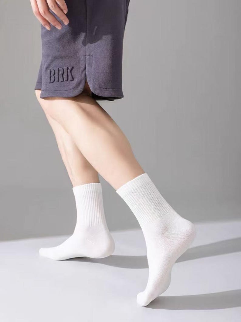 以下が含まれることがあります： 白いクルーソックスはグレーのショートパンツと合わせて着用されています。靴下はリブ編みで、ふくらはぎの中央まで届きます。ショートパンツの左脚には「BRK」の文字が刺繍されています。背景は無地のグレーです。
