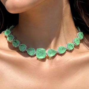 Può includere: Una collana color argento caratterizzata da una serie di pietre verde chiaro, traslucide, con taglio a goccia e a cuscino. Ogni pietra è circondata da un sottile bordo di piccoli cristalli chiari e scintillanti, creando un design a girocollo.