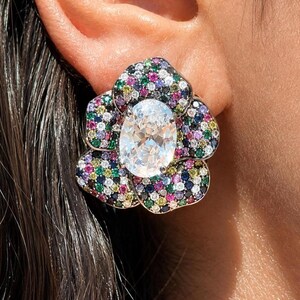 May include: A flower-shaped earring featuring a large, central oval-cut clear stone surrounded by a dense pavé setting of small, multi-colored gemstones including pink, blue, green, yellow, and white stones set in a metallic frame.