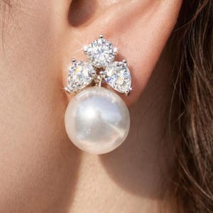 May include: A close-up of a white pearl earring with three sparkling diamond accents. The earring is attached to the ear. The pearl is large and round, and the diamonds are set in a cluster above the pearl.