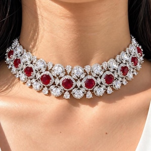 May include: A choker necklace featuring a series of deep red, cushion-cut stones arranged in a horizontal row. Each red stone is surrounded by a halo of small, clear, round-cut stones, with additional marquise and pear-cut clear stones in between.