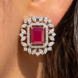 May include: A stud earring featuring a large, rectangular, deep red gemstone at the center, surrounded by a double halo of small, clear, marquise-cut and round-cut stones set in a silver-toned metal frame with a detailed, intricate border design.