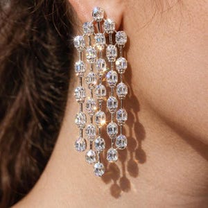 Puede incluir: Primer plano de un pendiente colgante brillante. El pendiente presenta múltiples piedras claras y facetadas dispuestas en un diseño en cascada. Las piedras están engastadas en un metal plateado, creando un aspecto glamuroso y elegante. El fondo está borroso.