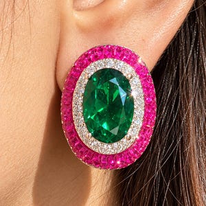May include: An oval-shaped earring featuring a large central green gemstone surrounded by a halo of small white stones and an outer border of vibrant pink square-cut stones. The piece is set in a metallic frame and worn on the earlobe.