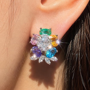 May include: A floral-shaped stud earring featuring a central oval clear gemstone surrounded by smaller oval gemstones in green, yellow, light blue, purple, and pink. The design includes small marquise-cut clear stones at the base, all set in silver.