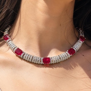 May include: A necklace featuring five rectangular red stones set at intervals. The segments between the red stones consist of multiple rows of small, clear, sparkling stones arranged in a cylindrical, band-like structure around the neck.