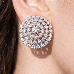 May include: A circular stud earring featuring a large, clear central stone encircled by many smaller, clear stones. The earring is set in gold-toned metal, creating a dazzling accessory. The earring is approximately 2.5 cm in diameter.