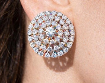 May include: A round, sparkling stud earring with a central, large clear stone surrounded by numerous smaller, clear stones. The earring is set in a gold-toned metal, creating a dazzling, eye-catching accessory. The earring is approximately 1 inch in diameter.