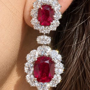 May include: A drop earring featuring two oval-cut deep red gemstones, each surrounded by a halo of small, clear, round-cut white stones. The two sections are connected by a small, rectangular, stone-encrusted link in a silver-toned metal setting.