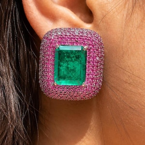 May include: A square-shaped earring featuring a large, rectangular emerald-cut green gemstone. The emerald is surrounded by a border of small, pink gemstones, set in a square shape. The earring is set against the skin.