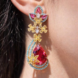 May include: A gold-toned earring with a floral design. The earring features a large, teardrop-shaped red gemstone, surrounded by blue and yellow accents. The top portion has pink and blue floral elements, with small white gems.