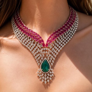May include: A wide, multi-layered necklace featuring a central pear-shaped green gemstone surrounded by small white stones. The upper section consists of two rows of rectangular red gemstones, while the lower section displays a lattice of white stones.