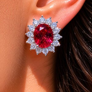 May include: A close-up of a dazzling stud earring. The earring showcases a large, oval-cut, ruby-red gemstone encircled by a halo of smaller, clear, round-cut diamonds. The setting appears to be silver or white gold, with the earring on an ear.