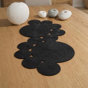 Könnte beinhalten: Ein Holztisch ist mit einer schwarzen, gewebten Tischset-Anordnung und dekorativen weißen und grauen Kerzenhaltern aus Stein gedeckt. Eine große weiße Vase steht ebenfalls auf dem Tisch. Der Raum hat eine neutrale Farbpalette.