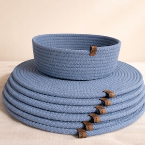 May include: A set of blue woven rope placemats and a matching basket. The placemats are stacked, with brown leather tags attached. The basket sits atop the placemats, creating a coordinated look for home decor.
