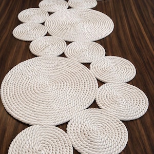 Può includere: Un set di tovagliette e sottobicchieri in corda bianca disposti su una superficie in legno scuro. Le tovagliette sono circolari, con un design a spirale. I sottobicchieri sono più piccoli e anch'essi circolari.