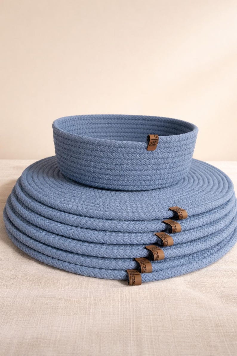 May include: A set of blue woven rope placemats and a matching basket. The placemats are stacked, with brown leather tags attached. The basket sits atop the placemats, creating a coordinated look for home decor.