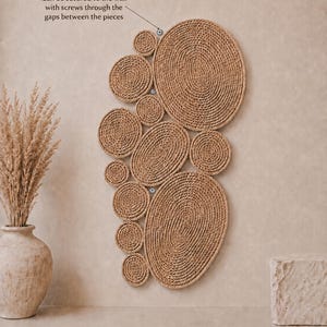 May include: A wall-mounted art piece composed of multiple circular and oval shapes made from coiled tan rope. The text states: Can be secured to the wall with screws through the gaps between the pieces. The design features varied sizes.