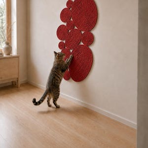 May include: A wall-mounted cat scratching post consisting of multiple circular discs of varying sizes arranged in a cluster. Each disc is constructed from coiled red rope, providing a textured surface for scratching. A tabby cat is shown interacting with it.