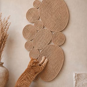 May include: A wall-mounted cat scratching post composed of multiple circular and oval discs made from coiled tan rope. An orange tabby cat is shown with its front paws extended, scratching the surface of the largest lower oval disc.