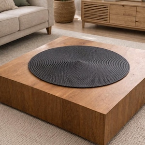 Può includere: Un sottopentola circolare grigio scuro in materiale tessuto è posto su un tavolino quadrato in legno. Il sottopentola ha un design a spirale. Il tavolo è in un soggiorno con un divano beige e un cesto intrecciato.