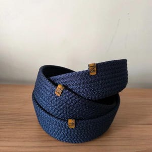 Puede incluir: Tres cinturones tejidos de color azul marino están apilados, creando un efecto espiral. Cada cinturón tiene una pequeña etiqueta rectangular de color marrón claro con la letra "N". Los cinturones están sobre una superficie de madera.