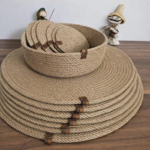 Może przedstawiać: Zestaw tkanych mat i podkładek z juty. Okrągłe maty i podkładki są uzupełnione małą miską, wszystko w gnieździe. Każdy element ma brązową sk&oacute;rzaną metkę. Zestaw znajduje się na drewnianej powierzchni.