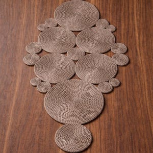Può includere: Runner da tavolo decorativo realizzato con corda marrone chiaro strettamente avvolta. Il runner presenta una serie di forme circolari e semicerchi disposti in un motivo simmetrico. Il runner è posto su una superficie di legno.
