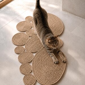 May include: A brown tabby cat stretches across a floor mat constructed from multiple tan rope coils of varying sizes. The mat features a large central oval shape surrounded by several smaller circular rope segments arranged in a cluster.