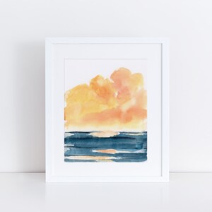 May include: A watercolor painting of a sunset over the ocean. The sky is a vibrant orange and yellow, and the water is a deep blue. The painting is framed in a white frame.