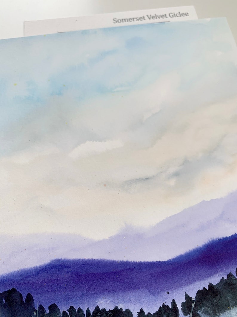 May include: Watercolor landscape art print featuring a serene mountain scene. The artwork showcases a gradient of colors, with a light blue sky, soft white clouds, and layers of purple mountains. The bottom of the print has dark trees. The text "Somerset Velvet Giclee" is visible.