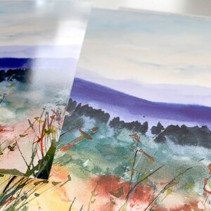 May include: Comparison of two types of paper for prints: Matte Photo and Somerset Velvet Giclee. The image shows a watercolor painting of a landscape with a blue sky, green grass, and red flowers.