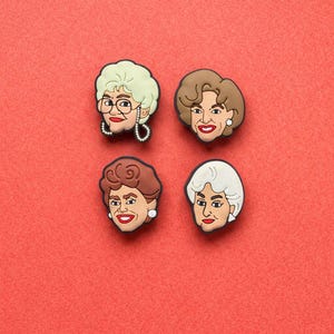 May include: Four cartoon face charms, each depicting a different woman with distinct hairstyles and facial features. The charms are set against a solid red background. The faces have different hair colours, including blonde, brown, and white.