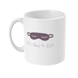 Op de afbeelding: Witte keramische mok met een paars slaapmasker ontwerp en de tekst "It's Okay to Rest." De mok heeft een gebogen handvat en is geschikt voor warme of koude dranken. Een eenvoudig en ontspannend ontwerp.