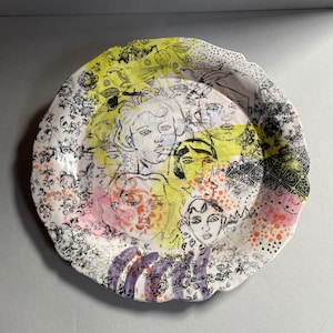 Könnte beinhalten: Ein weißer Keramikteller mit gewelltem Rand zeigt ein farbenfrohes Design aus überlappenden Gesichtern und floralen Mustern in Schwarz, Rosa, Gelb und Lila.