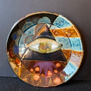 May include: A ceramic plate with a dark blue, brown, and white glaze. The center of the plate features a large, white eye inside a black triangle.