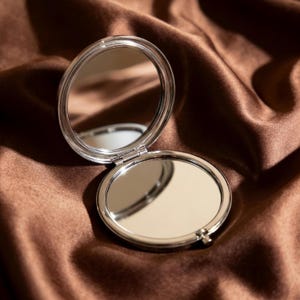 May include: A silver, round, open compact mirror with two reflective surfaces. The mirror is hinged and sits on a brown, silky fabric. The compact is open, revealing the mirrored surfaces.