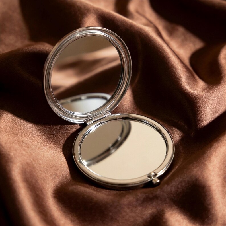 May include: A silver, round, open compact mirror with two reflective surfaces. The mirror is hinged and sits on a brown, silky fabric. The compact is open, revealing the mirrored surfaces.