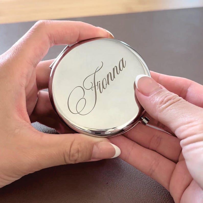 Personalized engraved compact mirror featuring a custom name in elegant script on a polished silver finish. Round pocket mirror shown held in hand, highlighting its sleek design and premium engraving. Perfect bridesmaid gift, maid of honor gift, bridal party favor, wedding keepsake, birthday gift, or personalized accessory for everyday use.
