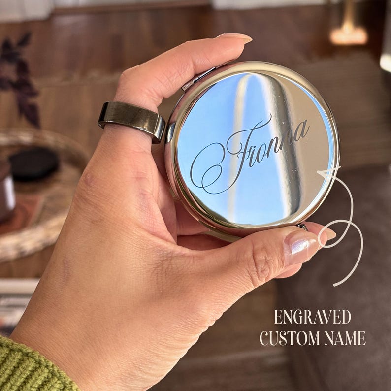 May include: A round, silver compact mirror with the engraved name "Fionna" in a cursive font. The text "ENGRAVED CUSTOM NAME" is below the mirror. The mirror is held in a hand.
