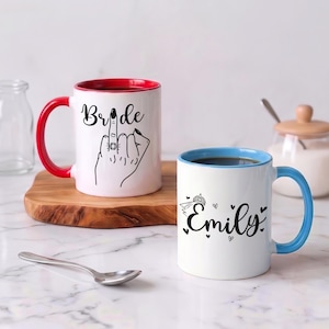 May include: Two white ceramic mugs with colored handles and rims. One mug has a red handle and rim, featuring the text Bride and a hand illustration with a ring. The other mug has a light blue handle and rim, featuring the text Emily and a tiara.