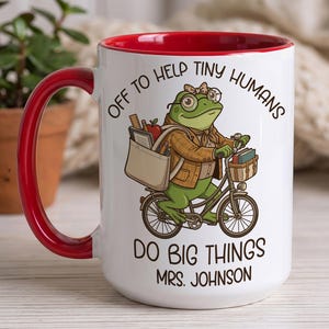 Könnte beinhalten: Ein weißer Keramikbecher mit rotem Innenraum und Henkel, verziert mit einer grünen Froschgrafik mit Brille und Schleife, der auf einem Fahrrad mit Schultaschen fährt. Der Text lautet: OFF TO HELP TINY HUMANS DO BIG THINGS MRS. JOHNSON.