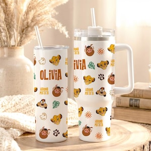 May include: Two white stainless steel tumblers featuring a repeating pattern of cartoon lion and warthog characters, paw prints, sun icons, and the text "Hakuna Matata" and "Olivia". One tumbler is slim with a straw, the other has a handle and lid.