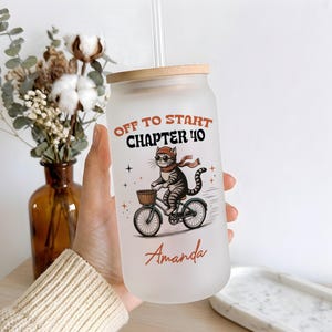 May include: A frosted glass tumbler with a bamboo lid and clear straw, featuring a graphic of a tabby cat wearing sunglasses and a scarf while riding a bicycle. The text reads "OFF TO START CHAPTER 40" in orange and black, with the name "Amanda" below.