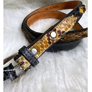 May include: A belt featuring a textured, snakeskin-patterned inlay in shades of tan, brown, and yellow, bordered by black leather edges. The belt includes silver-toned metal studs and a silver-toned buckle, with the text 2058-S 32 USA printed inside.