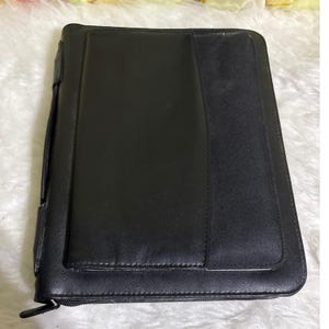 May include: A black rectangular zippered portfolio case featuring a smooth exterior finish with a vertical panel design. The case includes a side handle for carrying and a secure zipper closure that runs along the perimeter of the document holder.