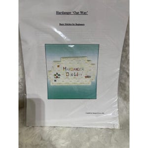 May include: A white instructional booklet titled Hardanger 'Our Way' Basic Stitches for Beginners, compiled by Margaret Peters, 2009. The cover features a central image of a white fabric piece with colorful embroidered text and small motifs.