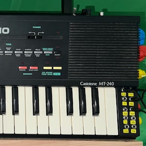 May include: A black Casio MT-240 keyboard with white and black keys. The keyboard features a green volume knob and various function buttons. The word "CASIO" is printed in white on the top. The keyboard has a handle.