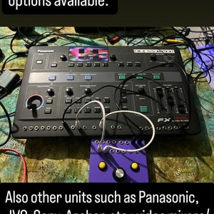 May include: A black Panasonic WJ-AVE7 digital video mixer featuring a small screen, numerous buttons, knobs, and sliders, connected via white cables to a small purple control box with two yellow buttons and a central silver knob.