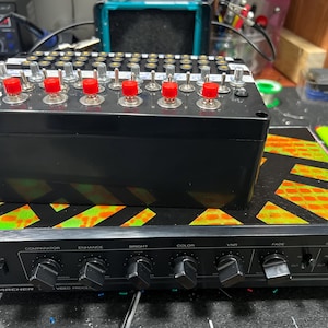 May include: A black rectangular electronic control box with multiple silver toggle switches and six red push buttons sits atop an Archer video processor. The processor features black knobs, a power switch, and a top surface with a neon orange and green pattern.